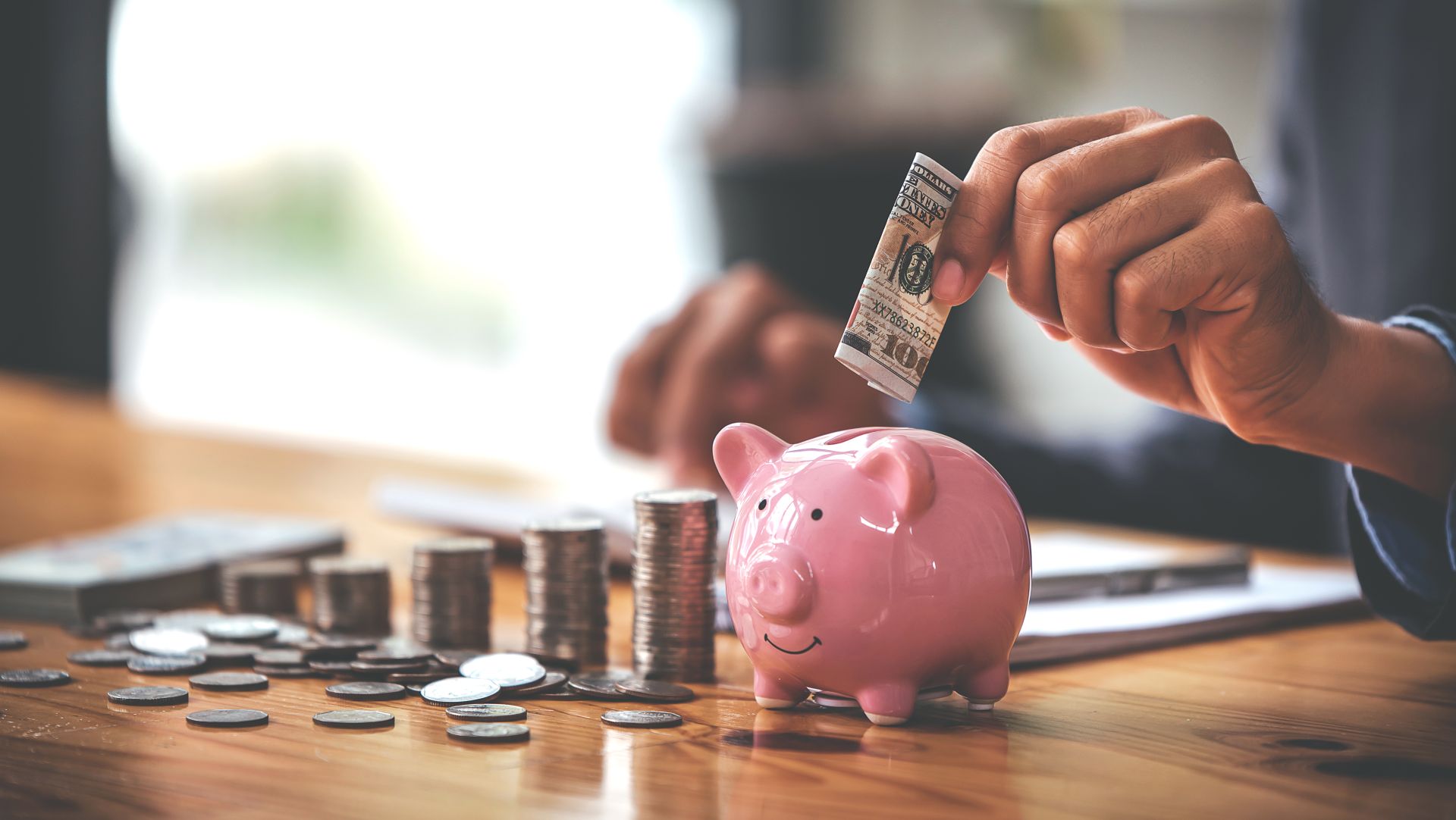 Person saving money by placing cash into a pink piggy bank with coins stacked nearby.