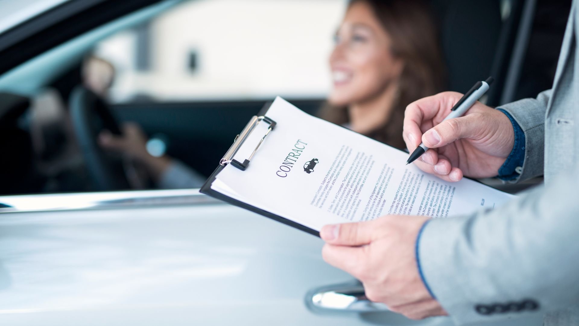 Person holding a car contract on a clipboard while a smiling woman sits in the driver’s seat of a car.