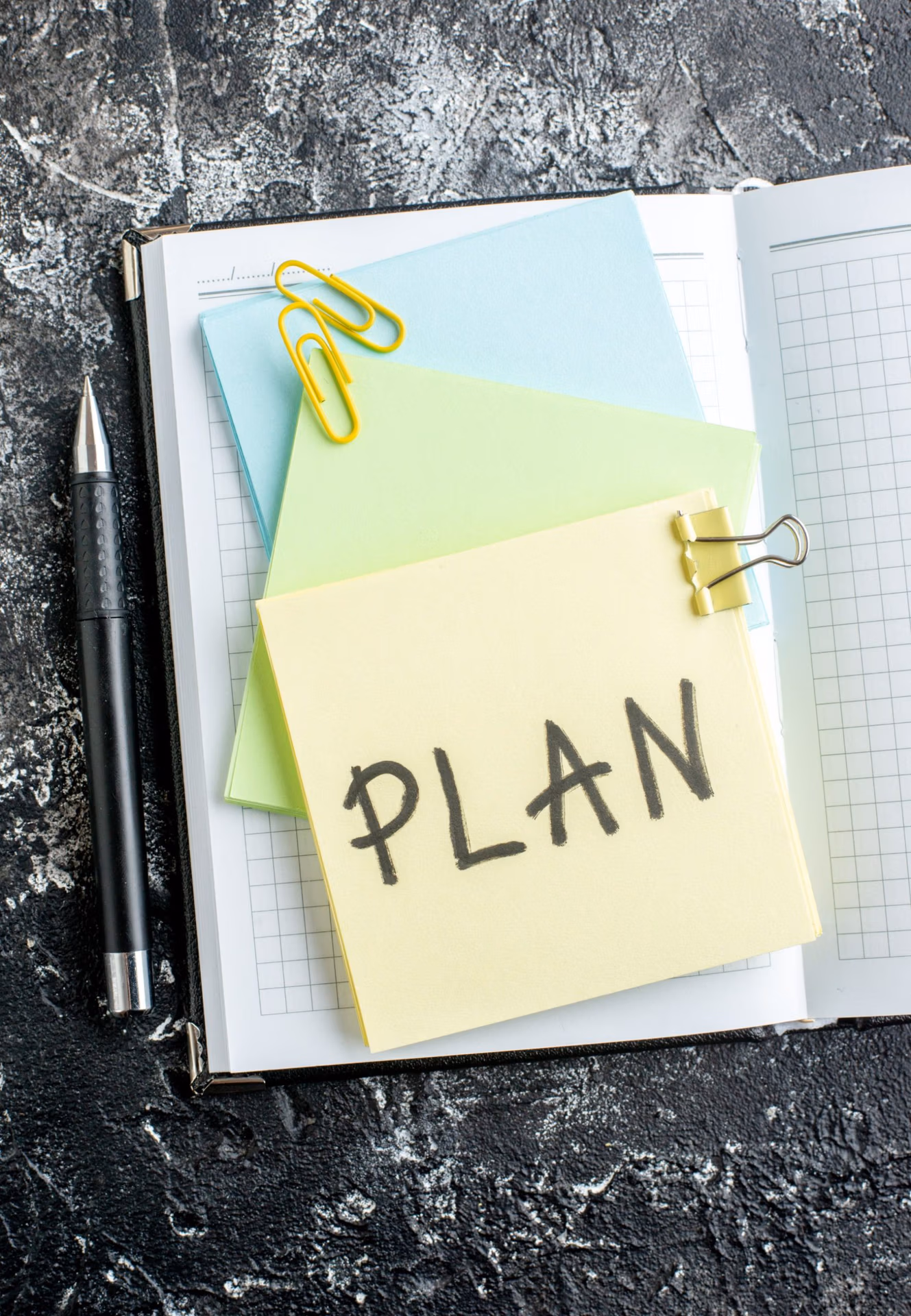 Notebook with sticky notes labeled “PLAN” clipped together, alongside a pen and paper clips.