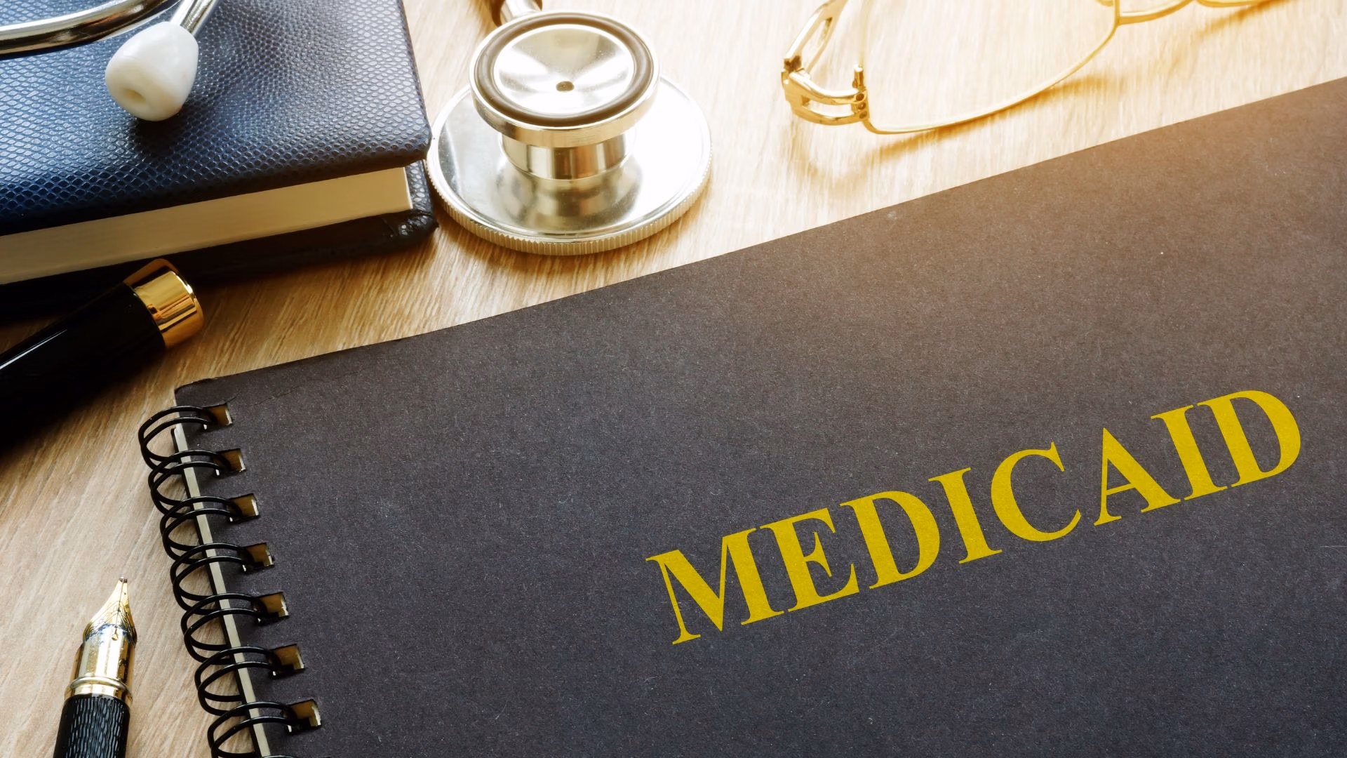 Black notebook labeled “MEDICAID” on a desk with a stethoscope, glasses, and a pen, symbolizing healthcare and eligibility planning.