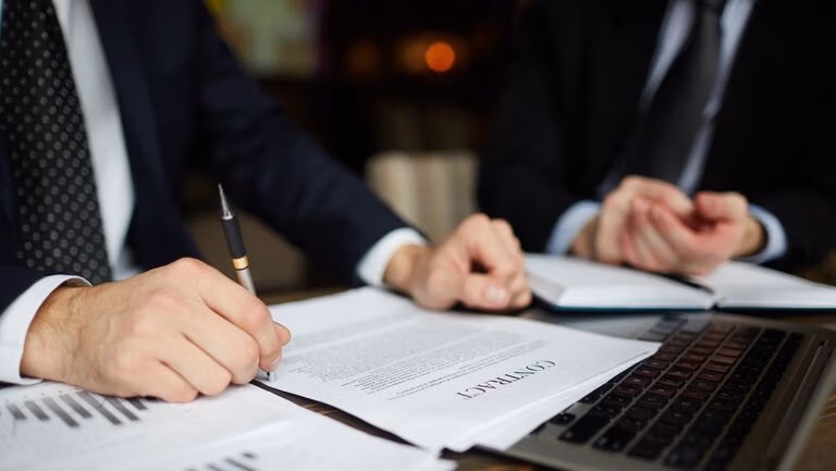 Business professionals reviewing and signing a legal contract at a desk with documents and laptop.
