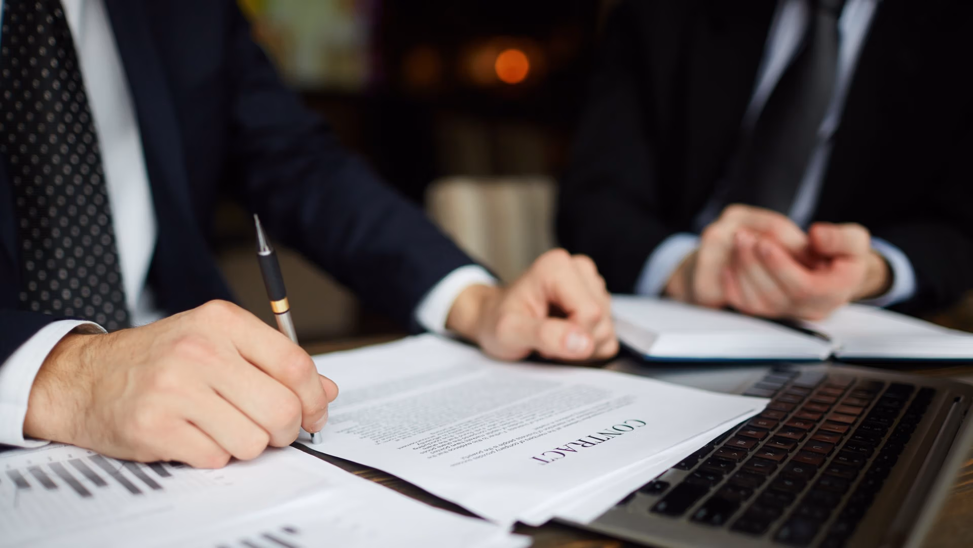 Business professionals reviewing and signing a legal contract at a desk with documents and laptop.