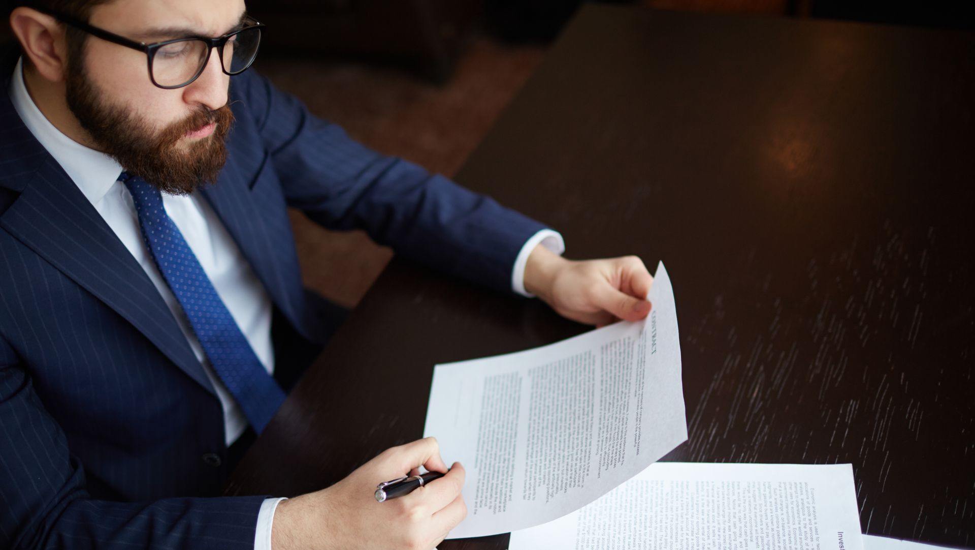 Man in suit carefully reviewing and holding legal contract documents at a desk.