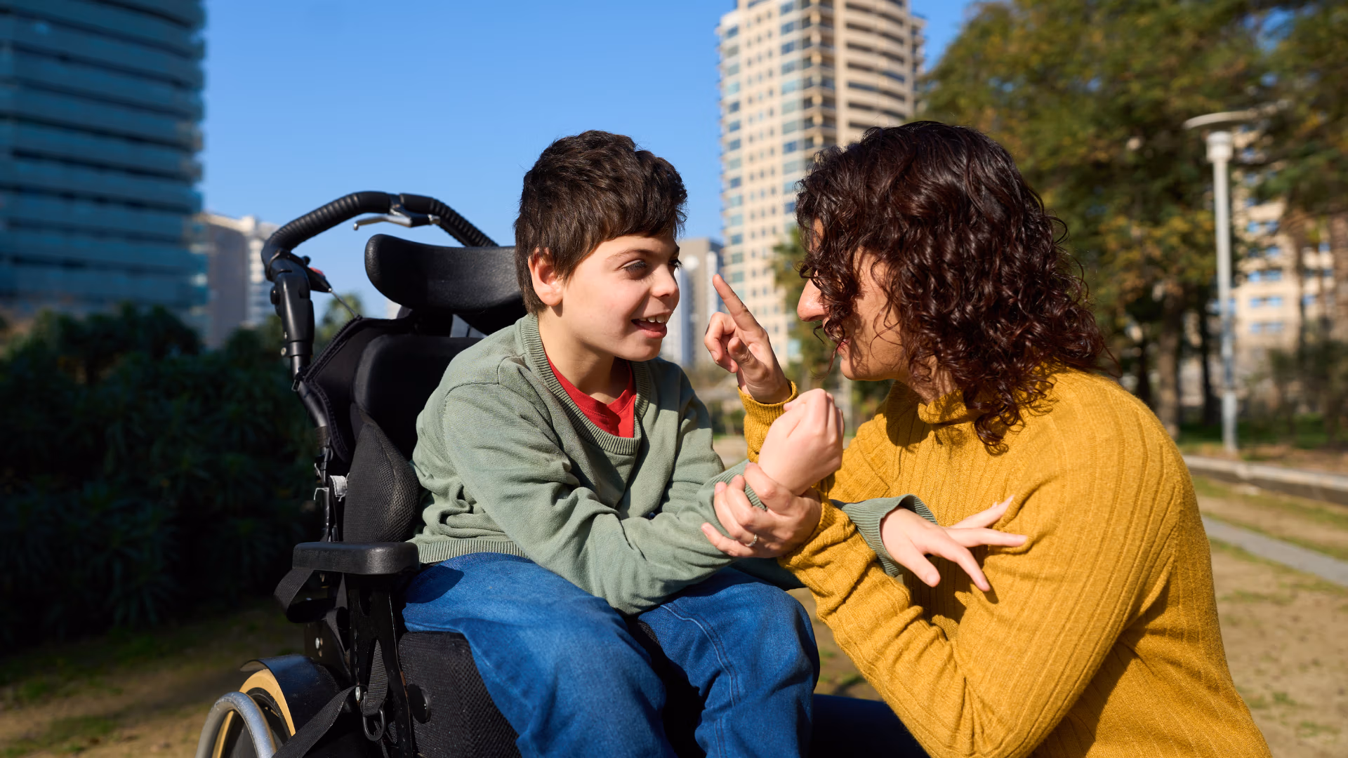 Mother and child in wheelchair smiling and communicating outdoors in the city.