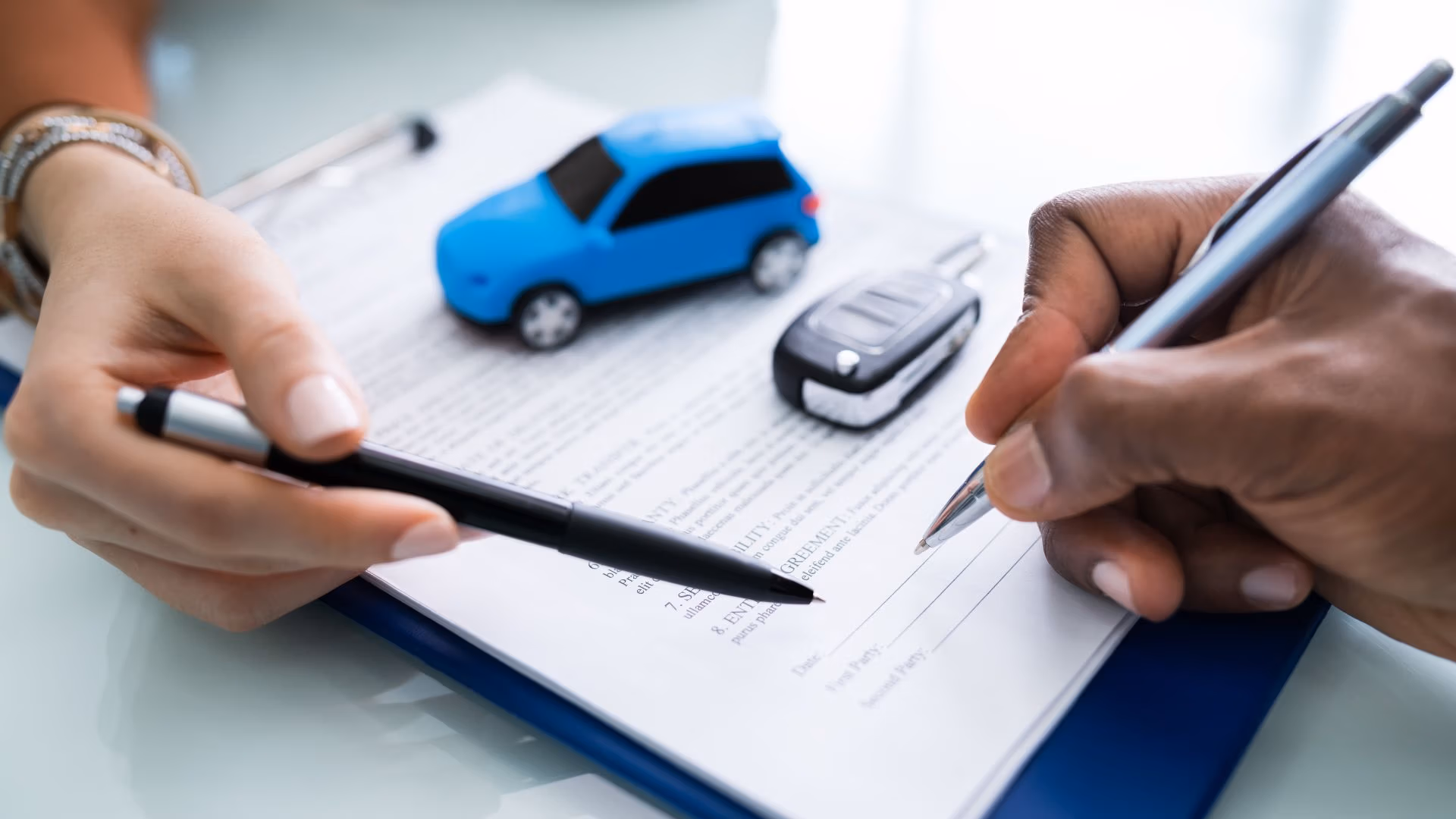 Person signing vehicle title paperwork with car key and toy car on the document.