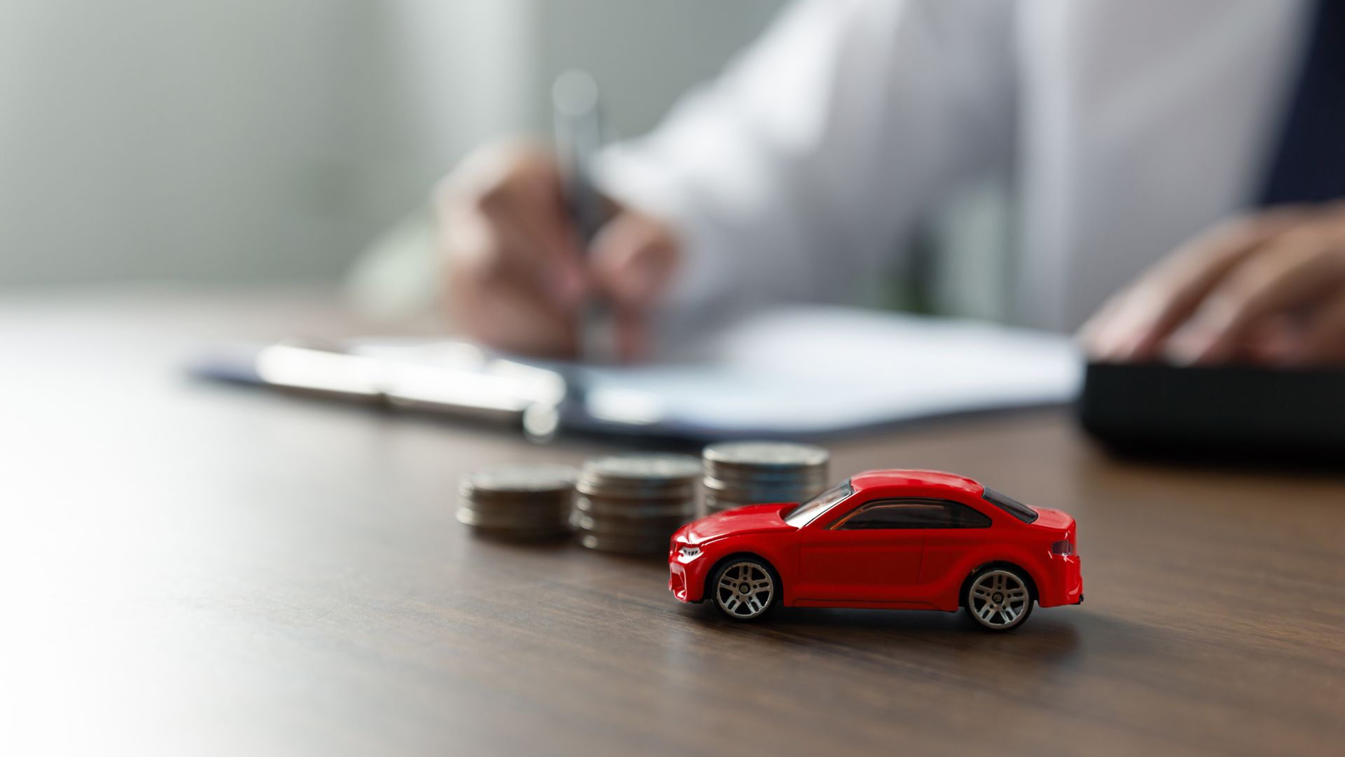 Red toy car beside stacked coins on a desk with a person signing documents in the background.
