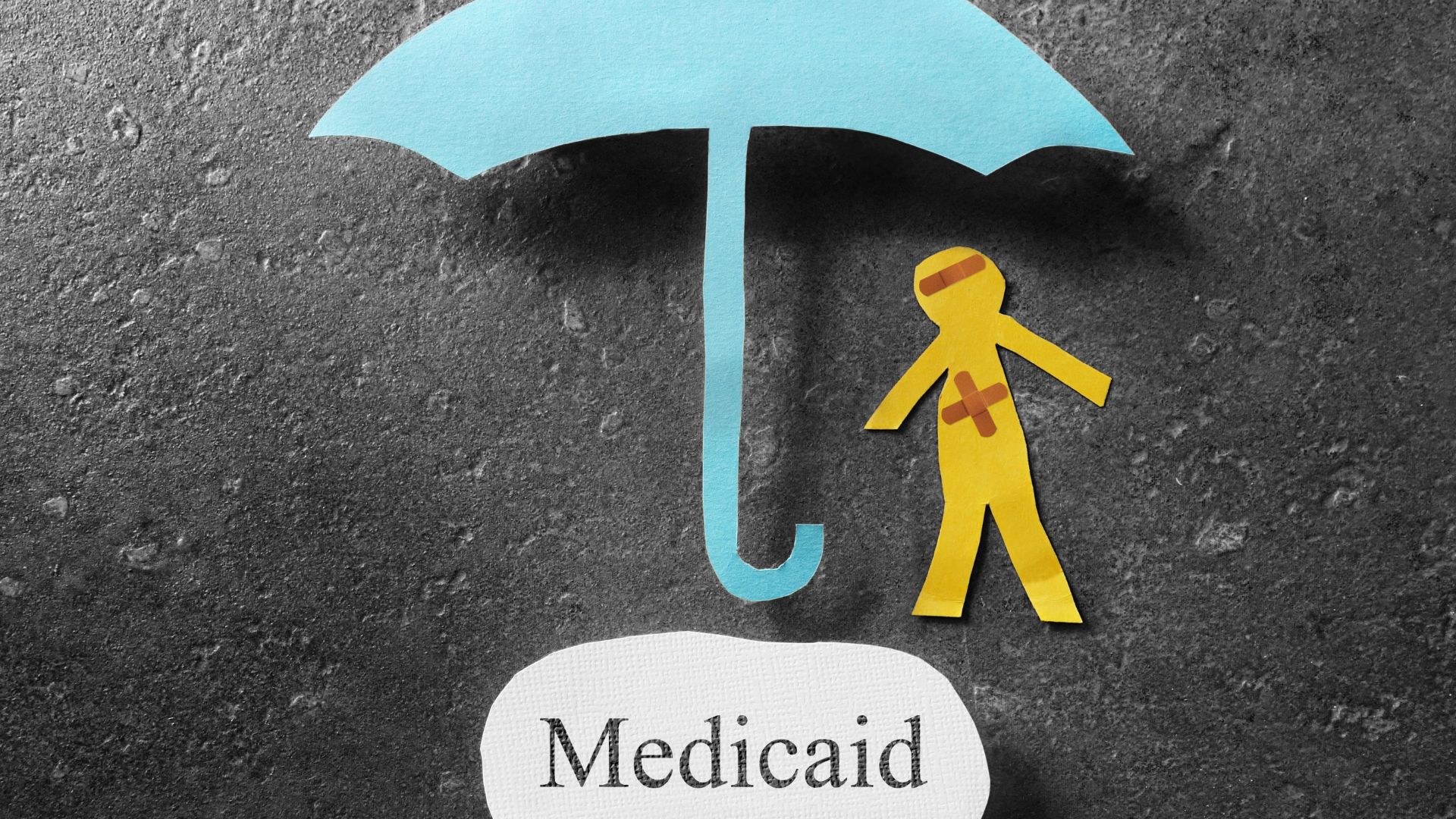 Paper cutout of an umbrella protecting an injured figure with the word “Medicaid,” symbolizing healthcare and coverage.