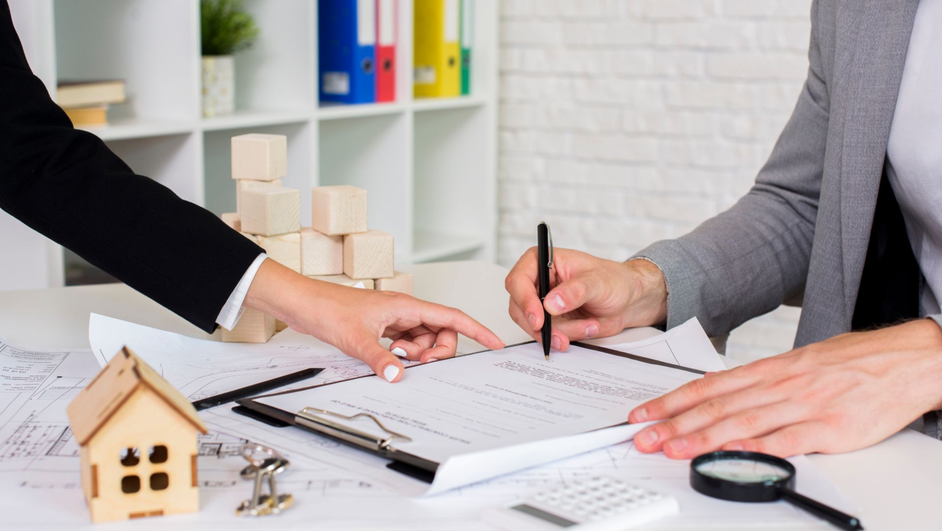 Close-up of people reviewing and signing legal real estate documents with a small house model and keys on the desk.