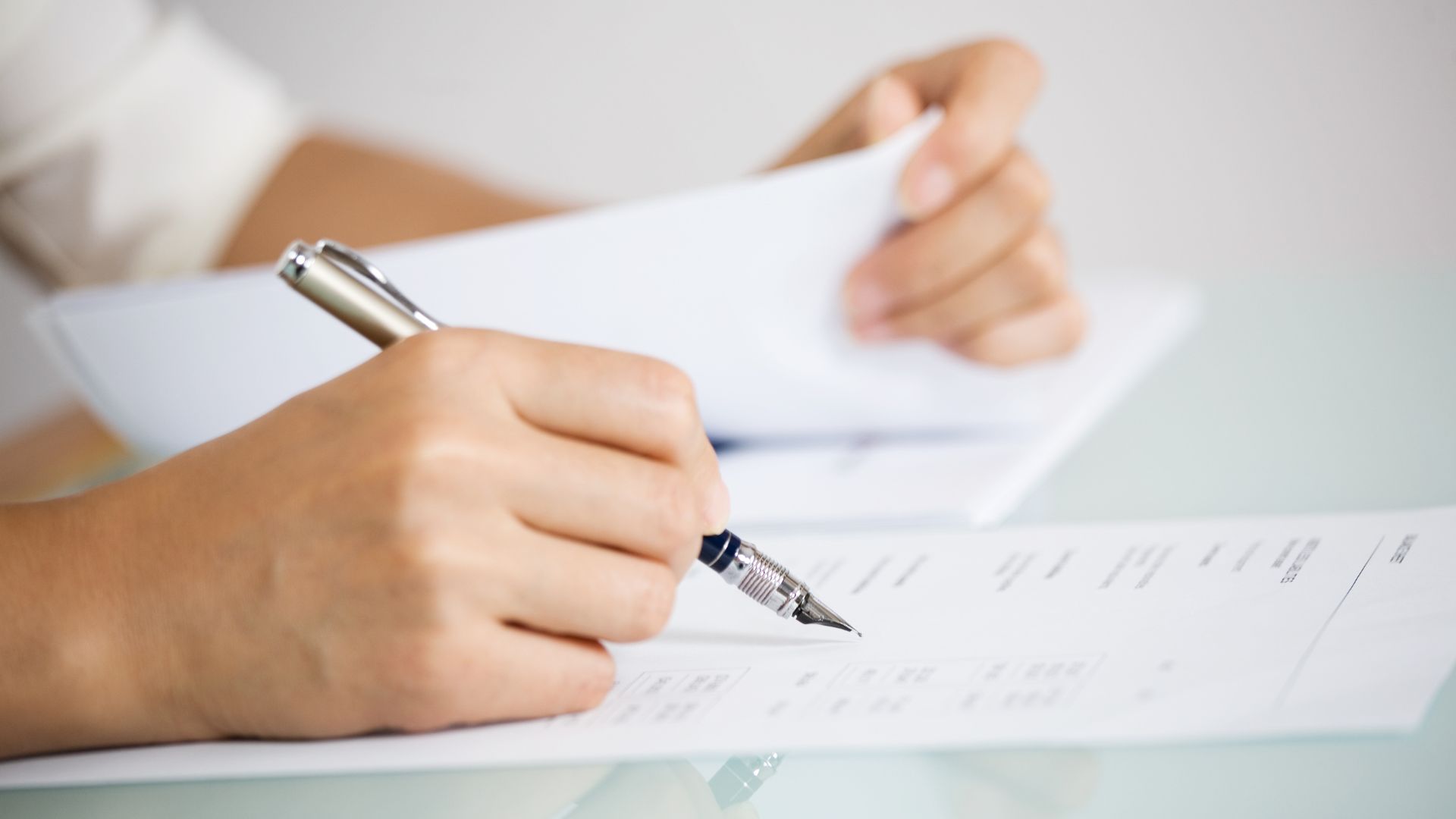 Close-up of hands reviewing and signing legal paperwork with a fountain pen on a glass desk.